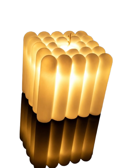 This image showcases a unique, sculptural decorative candle designed in a contemporary square format. The body of the candle is constructed from numerous vertical, cream-colored wax columns with rounded tops, creating a distinct ribbed or "bubble" texture around its entire perimeter. A single wick emerges from the center of the top grid. The candle is actively lit, emitting a warm, translucent golden glow that illuminates the wax from the inside out. It sits on a highly reflective, dark black surface, which perfectly mirrors the warm light and the pillared contours of the base.
Stylish cube-shaped LED light